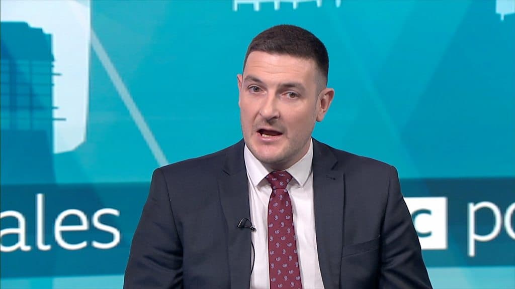 A cynical, dark oil painting style. A lonely, faceless politician in a grey suit sits in a small, leaky wooden boat labeled 'WELSH TORY' on a dark, choppy sea. In the distance, a larger ship is sinking, and a small, neon-orange buoy labeled 'REFORM' floats nearby. The sky is a bruised purple and grey, reflecting a sense of terminal decay and political hopelessness.