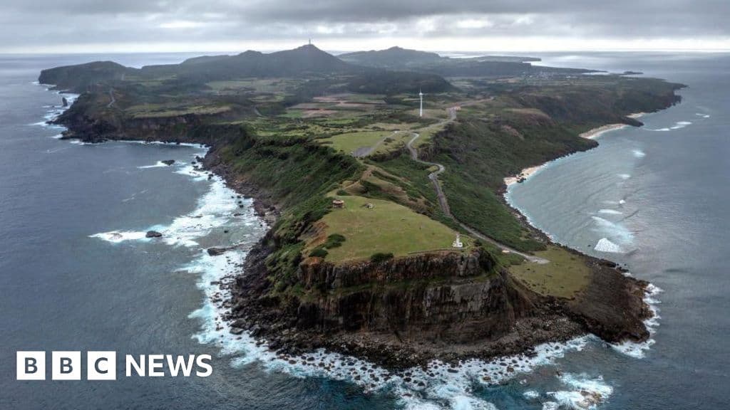 A moody, high-contrast editorial illustration showing a serene, lush green island in the ocean. In the foreground, out of focus, the cold steel silhouette of a military missile launcher looms. The sky is overcast and grey. Artistic style: Satirical political art, gritty realism.
