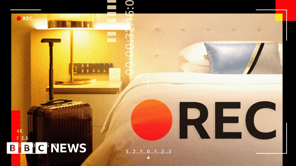 A close-up, gritty, low-light photo of a standard white electrical wall outlet in a dim hotel room. Inside one of the screw holes, a tiny, menacing glass camera lens glints slightly, reflecting a faint light. The texture is grainy and realistic.