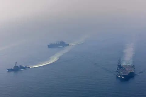 A hyper-realistic, cynical style editorial illustration showing a giant, gray toy aircraft carrier floating in a small, fragile porcelain teacup. The water in the teacup is turbulent and spilling over. On the side of the cup, a small 'Fragile' sticker is peeling off. The background is a dark, moody grey void. No text.