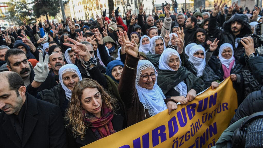 A cynical, wide-angle shot of a rainy Istanbul street at night. In the foreground, a pair of shiny black police boots stands over a dropped, mud-stained press ID card written in French. In the blurred background, the red lights of a police van glow through the mist, with the silhouettes of protesters and police in a chaotic struggle. The lighting is cold and oppressive, highlighting the futility of the scene.
