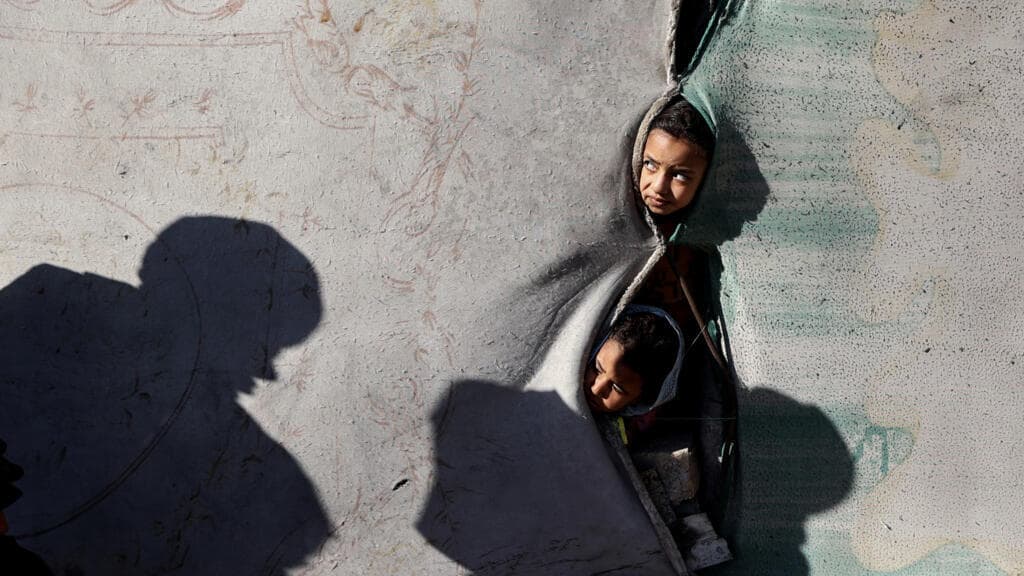 A surreal, cynical illustration of a desolate landscape in Gaza with crates of baby food stacked like a barricade. In the background, a golden, garish boardroom table labeled 'BOARD OF PEACE' floats in the sky, with silhouettes of suits clinking champagne glasses, ignoring the rubble below. High contrast, gritty style.