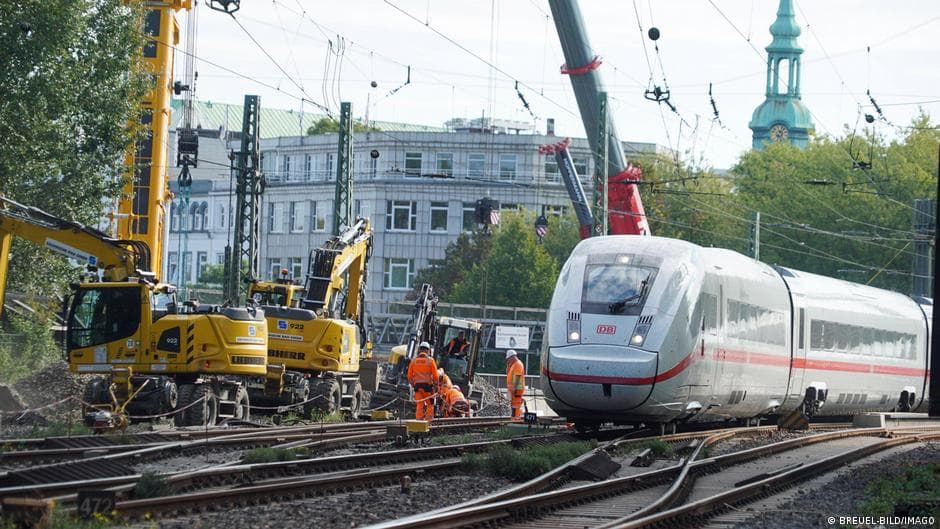 A hyper-realistic, satirical image of a sleek, high-tech German ICE train rusted and overgrown with weeds, stuck on a broken track in the middle of a desolate, grey wasteland. In the foreground, three politicians in suits (red, yellow, and green ties) are fighting over a single gold coin while ignoring a giant 'CANCELLED' sign on a flickering, digital departure board. The sky is a gloomy, industrial grey. Cinematic lighting, gritty detail.