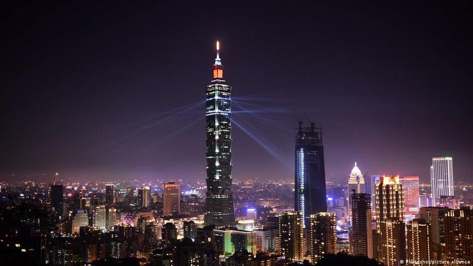A wide, cinematic shot of a lone climber, Alex Honnold, looking like a tiny ant against the massive, mirrored glass facade of a skyscraper. Below him, the city is a blur of neon lights. Reflected in the glass of the building is a giant, glowing red 'LIVE' button and the Netflix logo, with a faint shadow of a cheering crowd holding smartphones in the background.