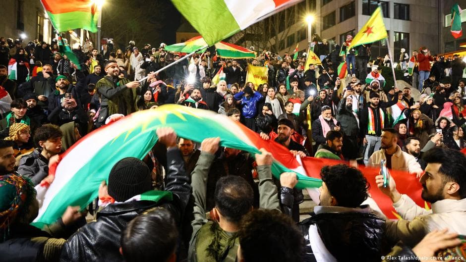 A high-contrast, satirical digital painting of a gray, rain-slicked German city square. In the foreground, a bored, well-dressed German bureaucrat in a gray suit checks his watch, completely ignoring a crowd of protesters behind him. The protesters are holding vibrant red, green, and yellow flags, but their faces are stylized as theatrical masks of exaggerated sorrow. In the background, the shadows of the German buildings morph into the silhouettes of tanks and ruined Syrian desert architecture, creating a jarring, surreal contrast between European comfort and Middle Eastern conflict.