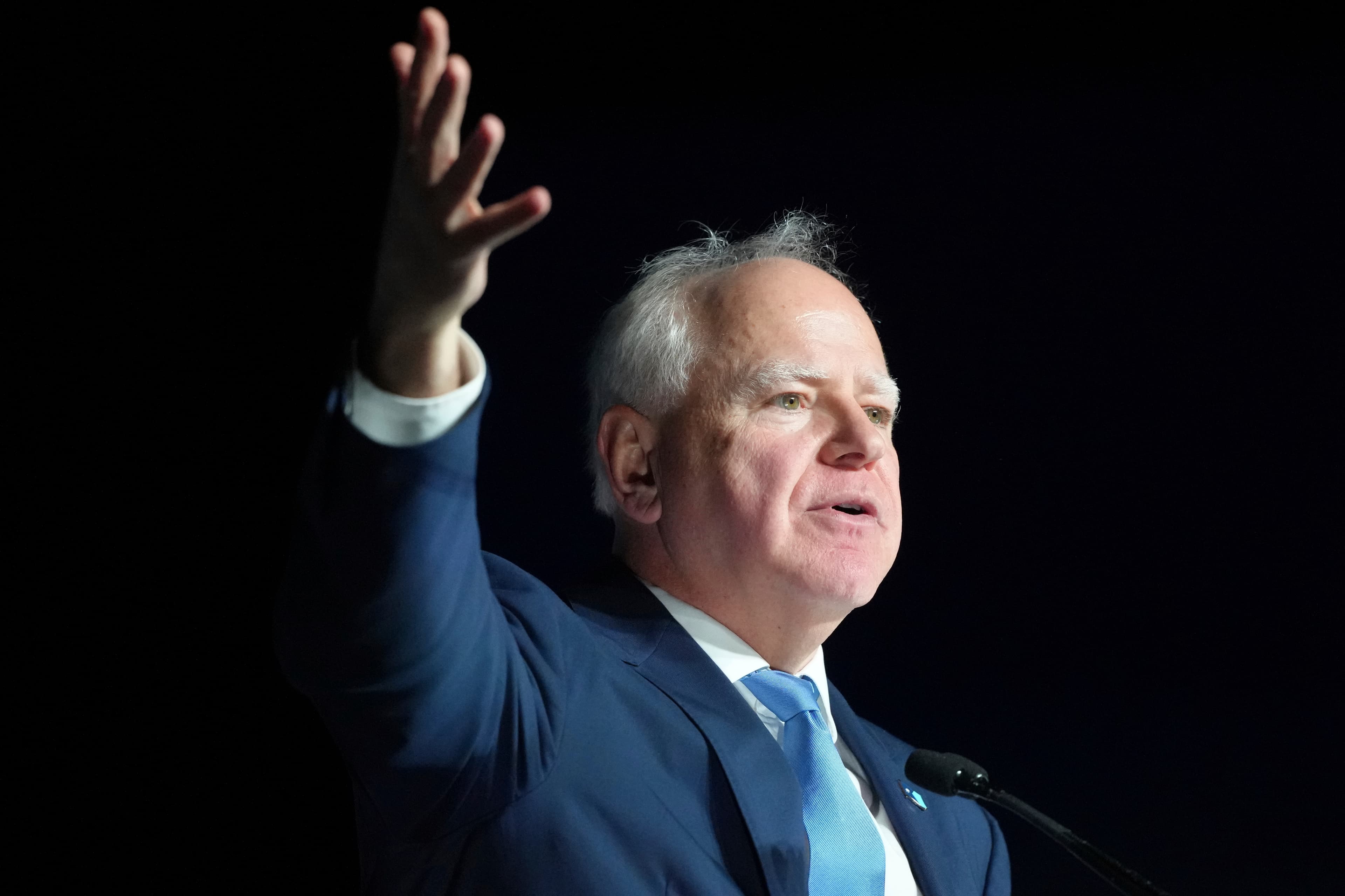 A satirical oil painting of Tim Walz walking away from a miniature Minnesota State Capitol building while carrying a suitcase labeled 'National Ambition', as Amy Klobuchar looms in the background holding a giant, menacing stapler like a scepter, set against a backdrop of a cold, grey Midwestern sky.
