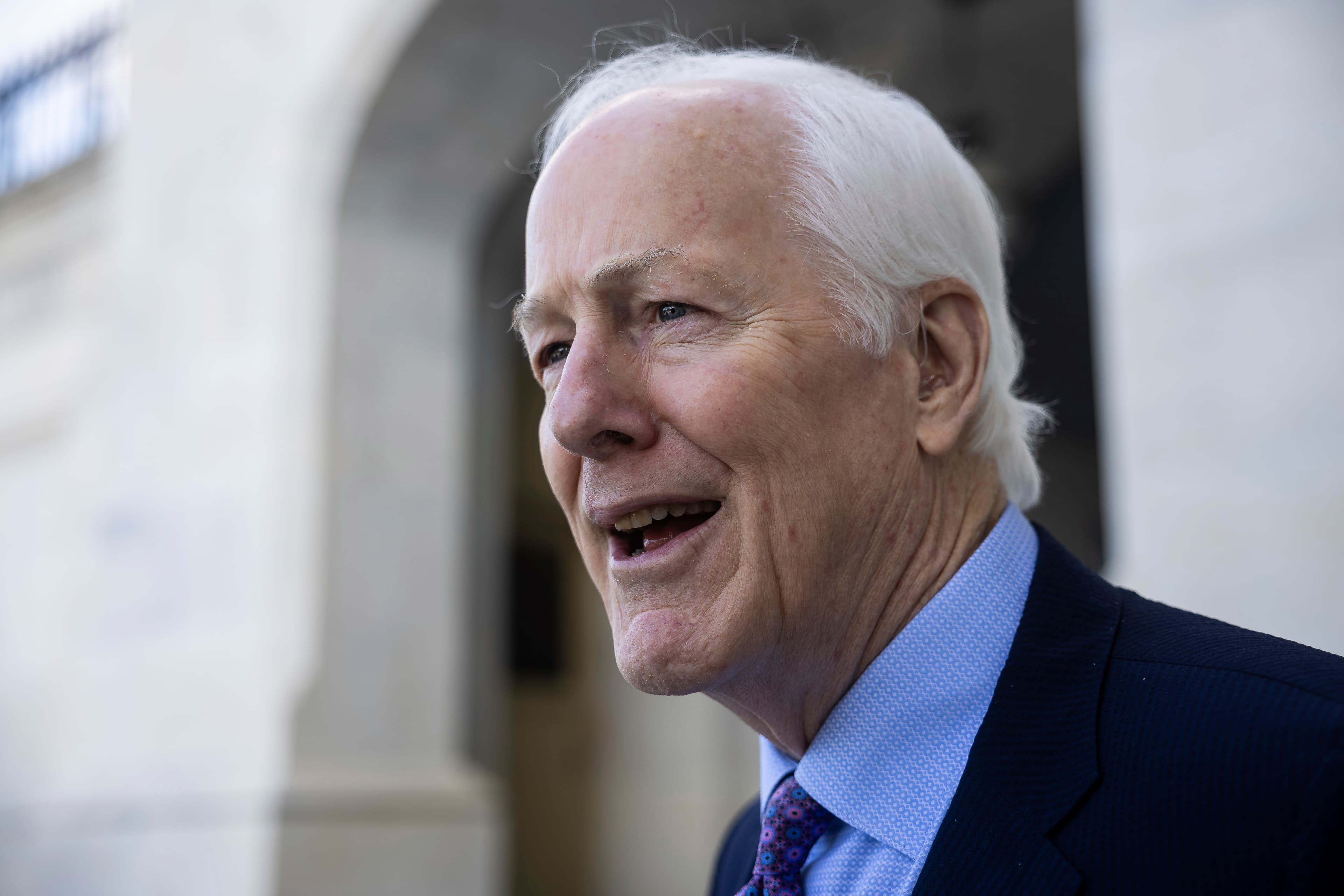A satirical oil painting of a dry, grey Senator John Cornyn sitting on a literal throne made of bundled hundred-dollar bills in the middle of a dusty Texas landscape, while faceless donor hands reach up from the soil to hand him more cash. High contrast, dark humor, editorial cartoon style.