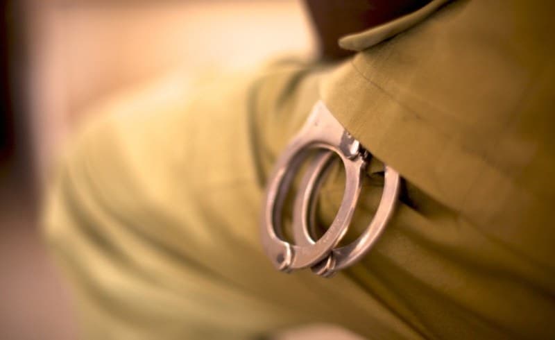 A gritty, dark satirical illustration. A high-ranking South African police official in an oversized fedora stands in front of a forest of microphones, his shadow on the wall looks like a circus clown. Behind him, a chalkboard displays the words 'ARRESTED' and 'QUESTIONED' with a giant, glowing neon question mark between them. The background shows a dimly lit, smoldering urban landscape. The art style is minimalist and sharp, using only charcoal grey, acid green, and deep red tones.