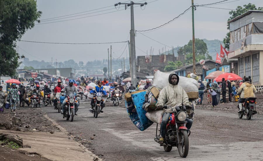 A gritty, hyper-realistic satirical illustration. A UN Blue Helmet stands in a desolate, war-torn Congolese landscape, looking through binoculars at a distant plume of smoke while standing on a massive pile of discarded human rights report papers. In the foreground, a luxury smartphone lies in the mud, its screen glowing with a 'low battery' warning. The lighting is harsh, cynical, and dramatic, emphasizing the contrast between the high-tech world and the primitive cycle of violence.
