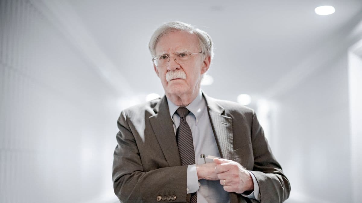 A satirical illustration of John Bolton with an oversized, sentient mustache that is whispering into his ear. He is standing on a mountain made of discarded war medals and television monitors, pointing a boney finger at a map of Ukraine while a orange sun sets in the background. The art style is gritty, dark caricature with heavy ink lines.