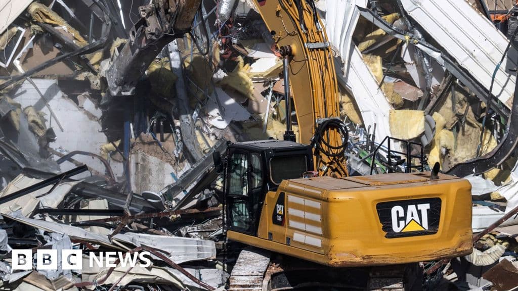 A hyper-realistic, gritty, wide-angle shot of a massive yellow industrial bulldozer clawing at the side of a white building with a faded blue 'UN' logo. Dust clouds fill the air, catching the harsh Mediterranean sunlight. In the foreground, a discarded, tattered UN flag lies in a puddle of muddy water. The background shows the ancient, stony skyline of Jerusalem under a pale, indifferent sky. High contrast, cinematic lighting, cynical atmosphere.