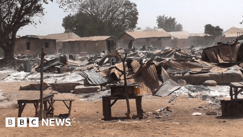 A wide, gritty photojournalistic shot of a dusty, arid landscape in Nigeria under a harsh, bright sun. In the foreground, freshly turned earth indicates a mass burial site with simple, crude wooden markers sticking out of the ground. The scene is desolate and empty of people, emphasizing silence and loss. High contrast, sombre tone.