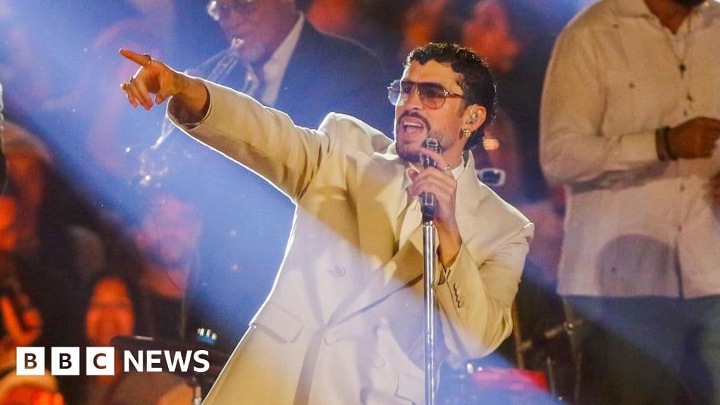 A hyper-realistic, cynical editorial illustration showing a tiny, lone figure of a singer standing on a massive, glowing American football field. The field is surrounded by towering walls of darkness. The stadium lights are blindingly bright but feel artificial and cold. In the background, vague shapes of corporate logos loom like giants. The mood is isolating and absurd, highlighting the smallness of the individual against a massive corporate machine.