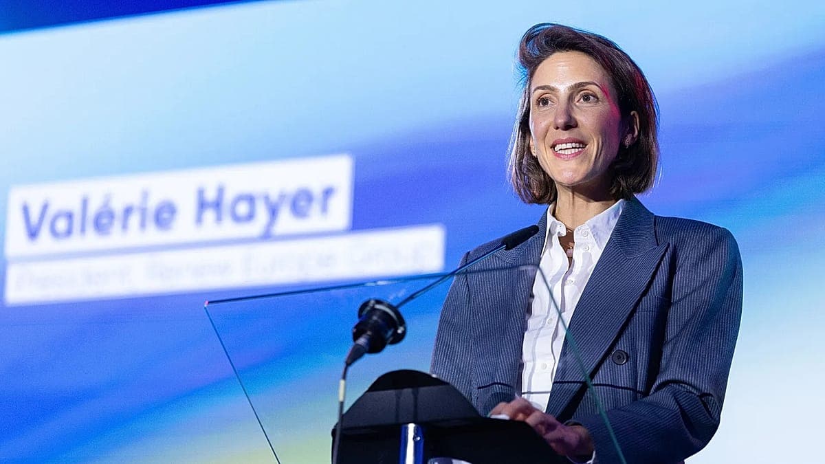 A sophisticated French woman in a sharp suit standing in a cold, brutalist Brussels office, holding a tiny, ornamental silver shield while a giant shadow of a man in a red tie looms over a map of Greenland in the background. Cinematic lighting, cynical atmosphere, 8k, hyper-realistic.