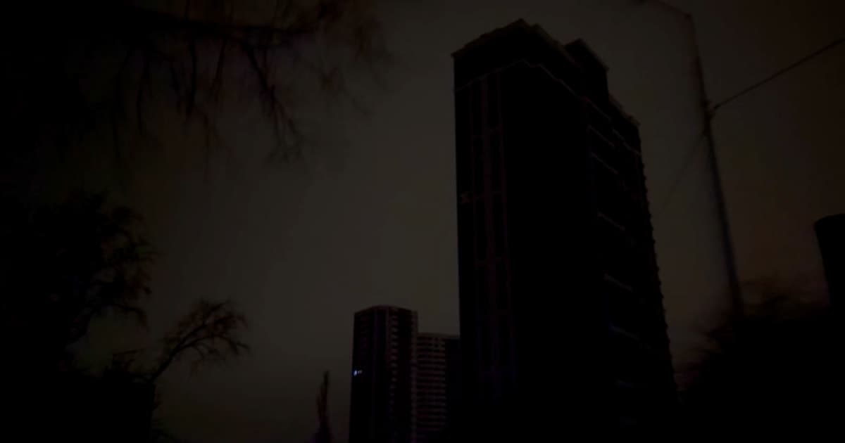 A gritty, noir-style illustration of a modern city skyline completely blacked out at night, covered in snow. Only a single, faint candlelight is visible in one window. The atmosphere is cold, blue, and desolate. No people visible, just the dark, freezing buildings.
