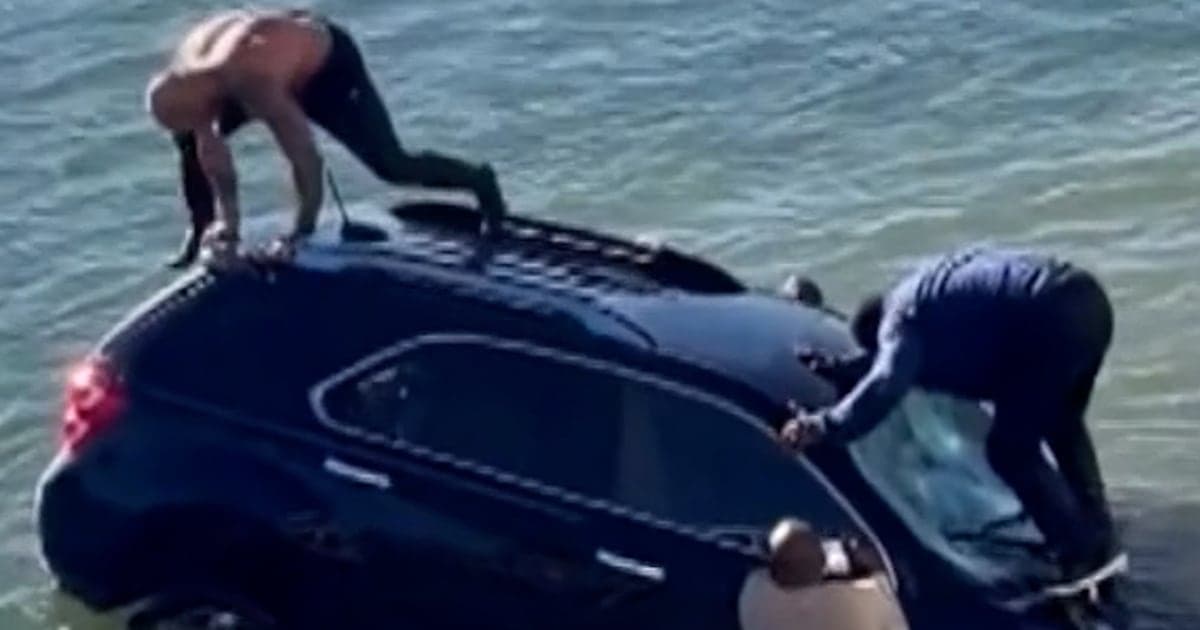 A hyper-realistic, gritty cinematic shot from a low angle of a dark silver SUV half-submerged in churning, cold grey ocean water next to a concrete boat ramp. A Navy rescue swimmer in a black wetsuit and yellow helmet is battling heavy spray to reach the driver's side window. The sky is a flat, oppressive grey, and the overall mood is one of bleak, industrial-scale rescue and human failure.