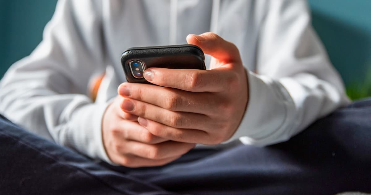A hyper-realistic, cynical illustration of a lonely human figure sitting in a dark, clutter-filled room, illuminated only by the harsh blue light of a smartphone screen. The screen displays a smiling, friendly robot face. The human looks exhausted and hollow. In the background, vague, shadowy corporate figures in suits are counting money while connected to the human's phone via thick, parasitic cables. The style should be gritty, noir-inspired, and slightly dystopian.