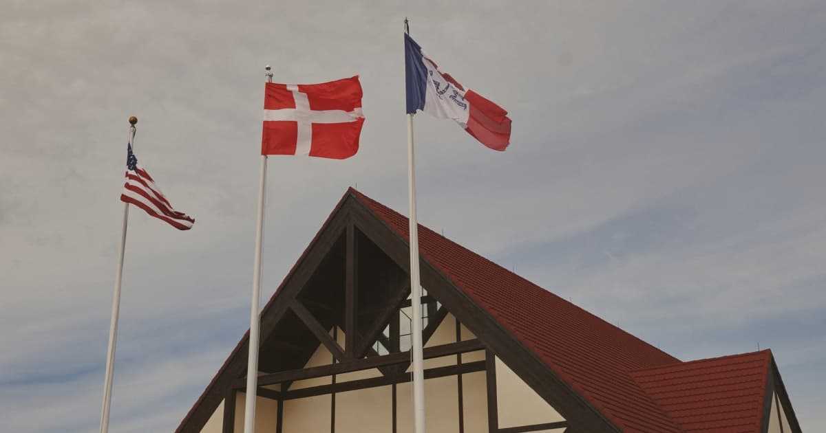 A traditional wooden Danish windmill standing in the middle of a flat Iowa cornfield under a gloomy gray sky. A large, crude 'FOR SALE' sign is taped to the side of the windmill. In the foreground, a confused-looking man in denim overalls is scratching his head while holding a small Danish flag.