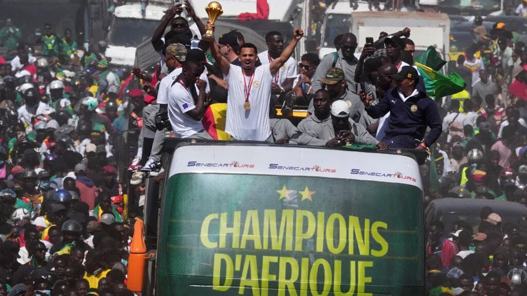 A gritty, high-contrast photograph of a massive crowd in Dakar, Senegal, viewed from a high angle. A bright gold bus is barely visible, engulfed by a sea of people in green and yellow. The air is thick with dust and heat haze. In the distance, a government building stands cold and grey, contrasting with the chaotic, colorful frenzy of the masses below. The lighting is harsh and cynical.
