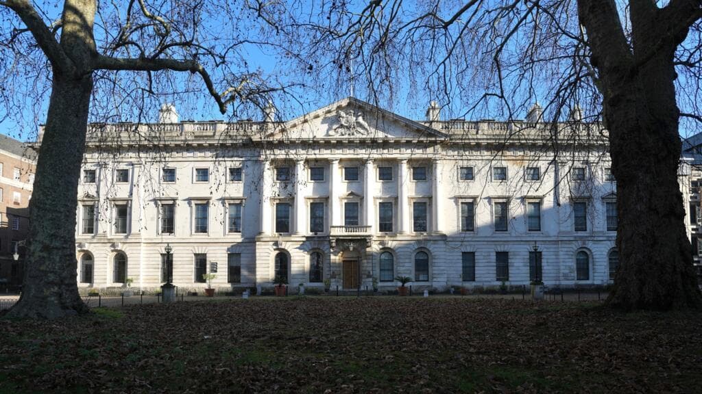 A satirical, dark-toned illustration of a massive, futuristic, brutalist fortress with Chinese architectural elements being constructed on the site of the historic Royal Mint Court in London. In the foreground, a group of tired-looking British politicians in suits are holding a 'FOR SALE' sign and a tray of tea, bowing toward the building. The sky is a smoggy, polluted grey, and the Tower of London in the background looks small and decrepit in comparison to the towering, high-tech embassy. Sharp, cynical art style, high contrast.