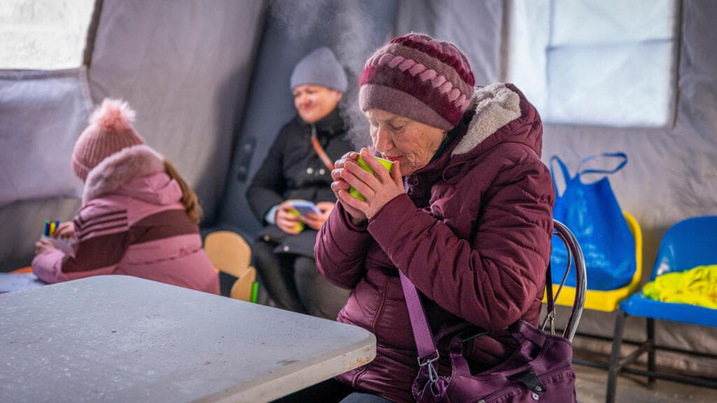 A gloomy, realistic oil painting of a modern high-rise apartment building in Kyiv at night. The building is completely dark and silhouetted against a snowy, dark blue sky, except for a single window illuminated by a weak, flickering candle. Snow is falling heavily. In the distance, faint smoke rises from the horizon. The mood is cold, lonely, and stark.