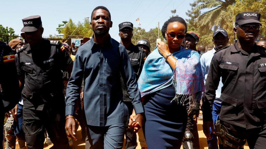 A cynical, dark-humored illustration of a Ugandan political landscape. On one side, an aging dictator in military fatigues sits on a throne made of ballot boxes, handing a golden rifle to his son. On the other side, a man in a red beret hides behind a large, cracked musical note. In the background, the word 'DEMOCRACY' is being erased from a chalkboard by a soldier. The style is sharp, editorial, and acid-etched.