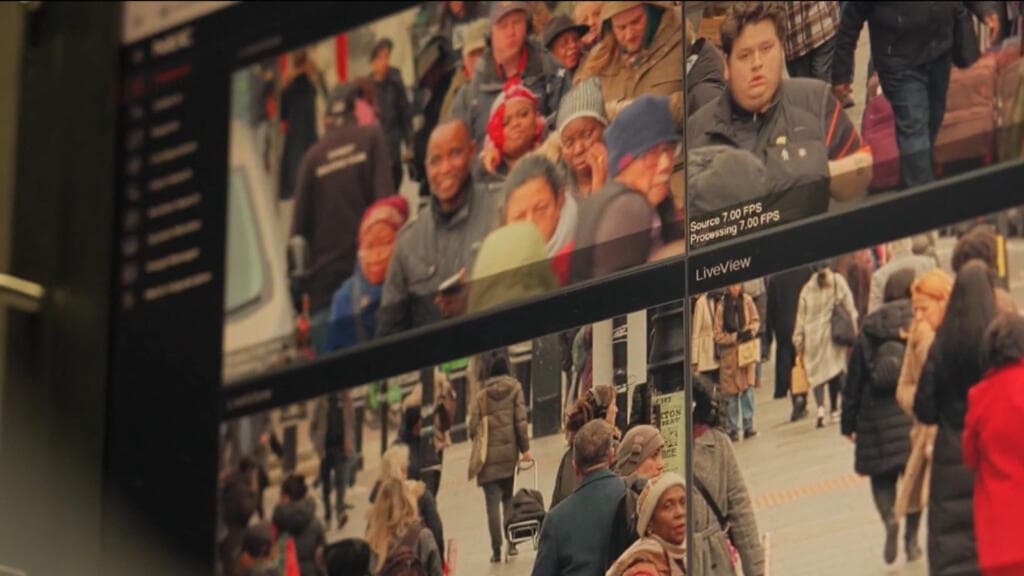 A gritty, grey London street scene in a satirical style. The buildings are covered in hundreds of oversized, realistic eyeballs and CCTV cameras twisted together like vines. Below, faceless commuters in grey suits walk with barcodes on their foreheads. The atmosphere is rainy, gloomy, and oppressive, with a single bright spotlight shining down from a police drone.