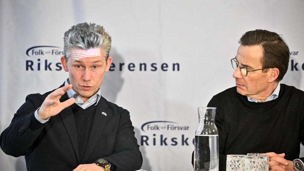A satirical political cartoon showing a giant, orange-tinted hand holding a massive 'FOR SALE' sign over a map of the Arctic, while a small, neatly dressed Swedish bureaucrat shakes a tiny finger at it. In the background, a German official is holding a paper shield labeled 'SUPPORT,' which is clearly made of cardboard. The ice beneath them is cracking, and the sky is filled with the logos of various multinational corporations.