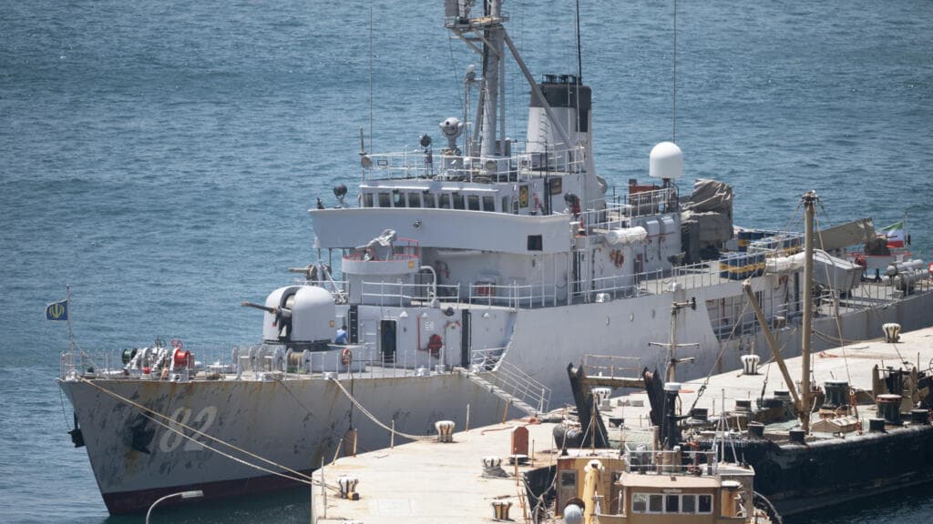 A satirical political cartoon showing a confused South African admiral in a bathtub with a toy Iranian warship, while a furious Uncle Sam watches through a window and a South African politician hides a 'Do Not Invite Iran' memo behind her back. High contrast, cynical caricature style.