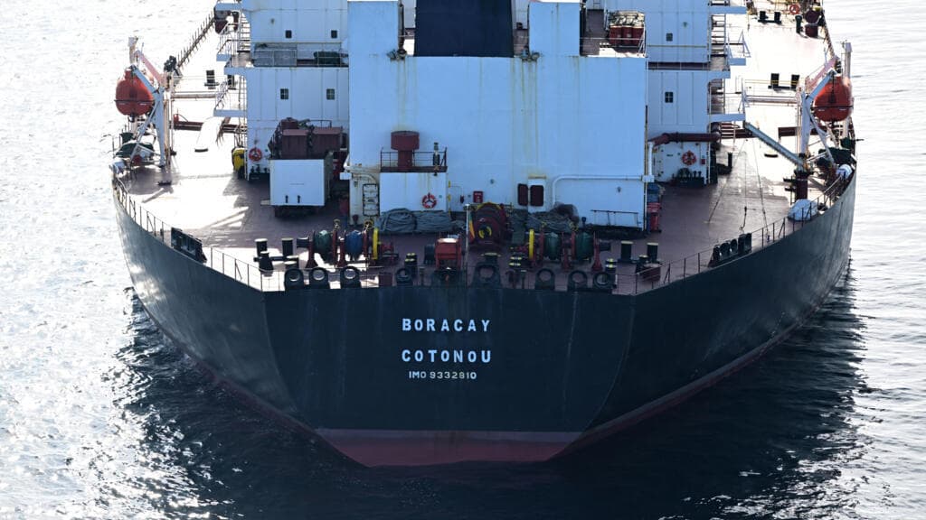 A gritty, satirical oil painting of a rusted, crumbling oil tanker with ten different mismatched national flags flying from its mast, sailing through a sea of thick black ink under a dark, stormy sky. On a distant, pristine shore, a group of portly bureaucrats in suits are standing in a circle, holding a large paper banner that says 'SANCTIONS' while they all have their eyes tightly shut and fingers in their ears.