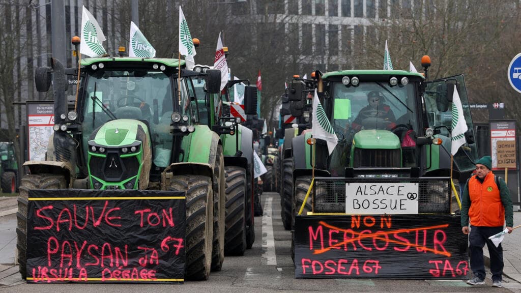 A cynical, high-contrast photograph of shiny, modern green tractors blockading the curved glass-and-steel European Parliament building in Strasbourg. The sky is a grim, overcast grey. In the foreground, a pile of manure sits on the pristine cobblestones. The reflection of the tractors in the glass of the parliament building creates a distorted, chaotic image of modern European bureaucracy and agricultural protest.
