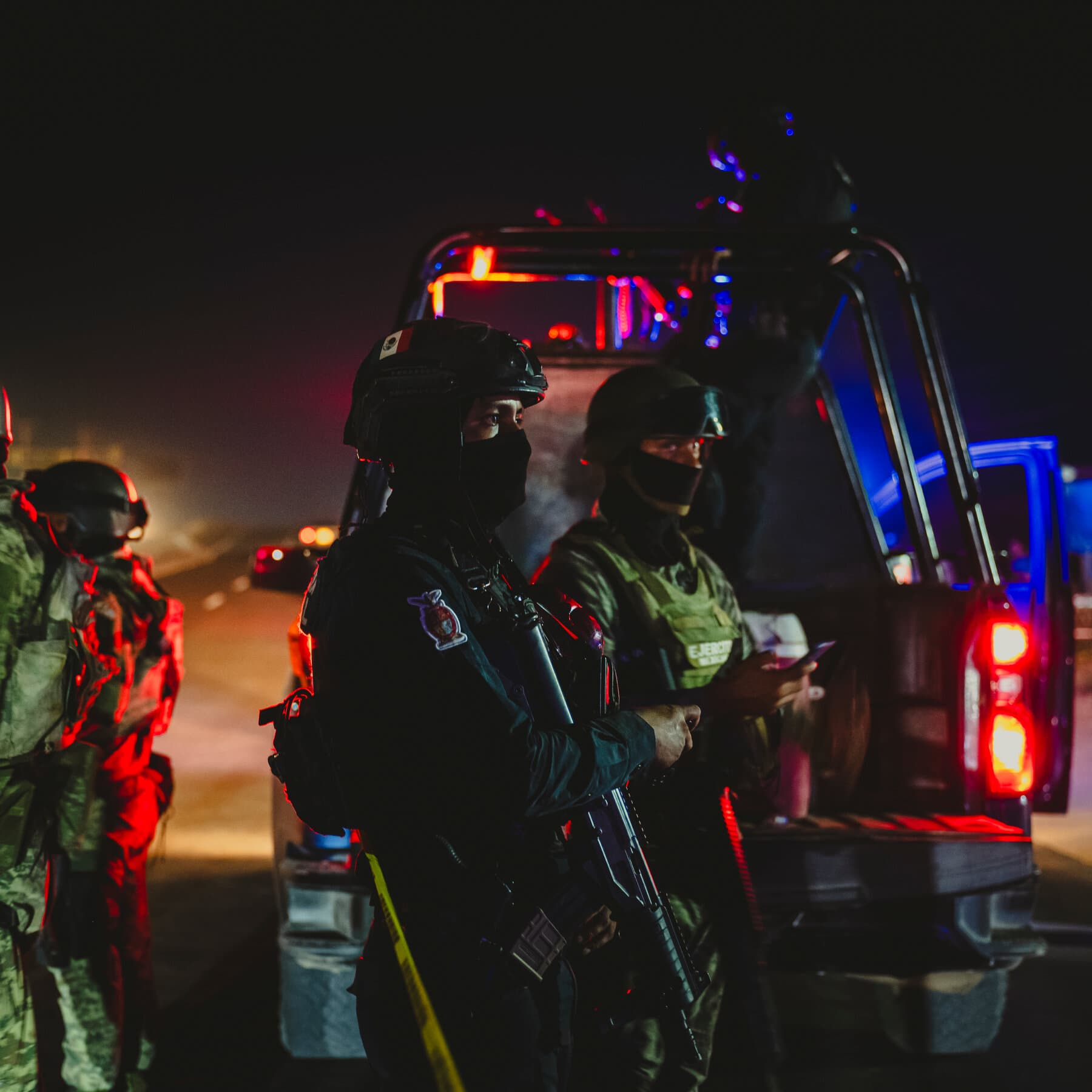 A cynical, dark satirical illustration in the style of a gritty political cartoon. In the foreground, a large gift-wrapped box labeled '37 Sacrifices' sits on the U.S.-Mexico border. In the background, a silhouette of a drone shaped like a screaming face hovers over a Mexican landscape. On the U.S. side, a gold-plated podium stands empty. The colors are muted, sickly yellows and deep shadows, emphasizing a sense of existential dread and political farce.