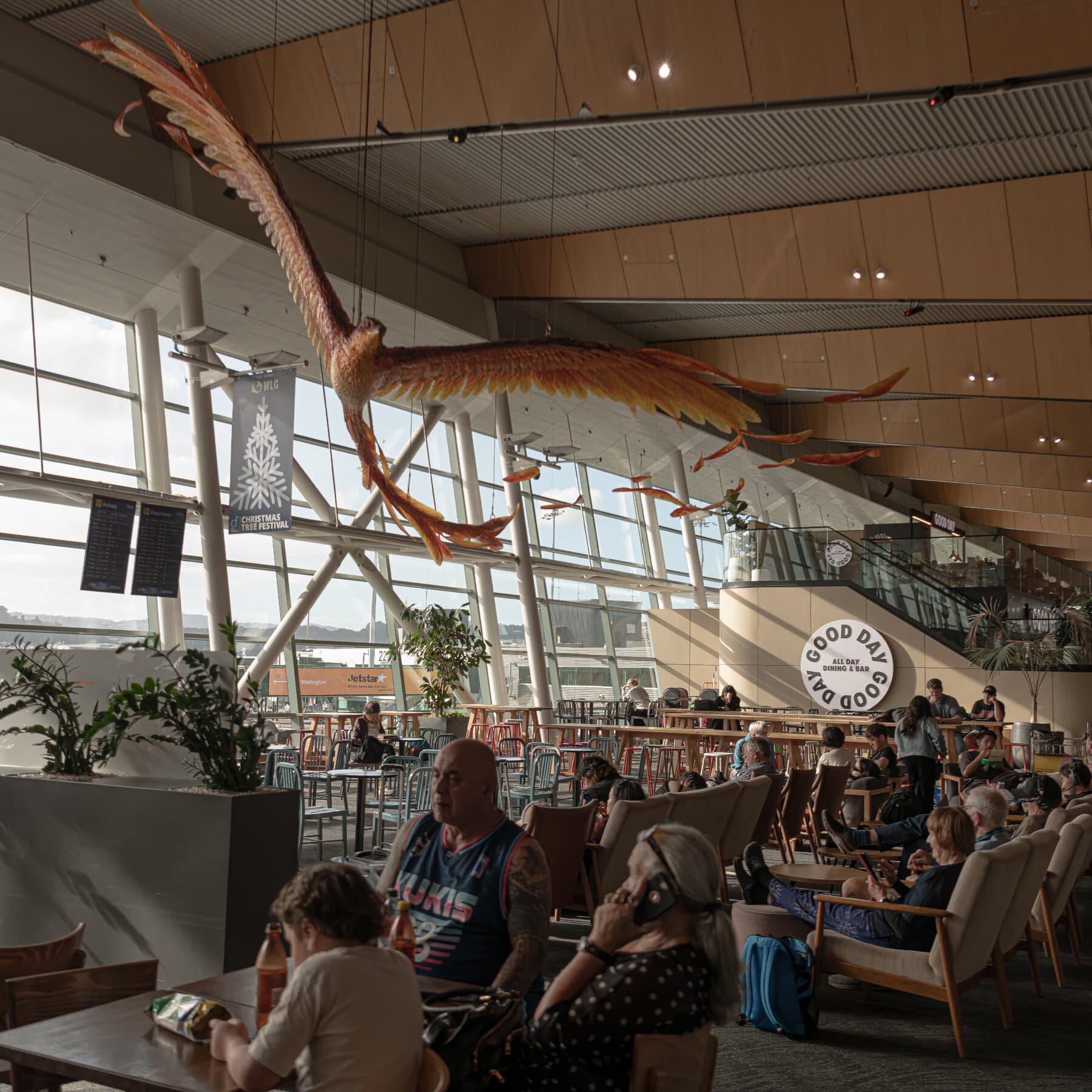A satirical, bleak illustration of a stream of generic, tired-looking people (New Zealanders) carrying suitcases, walking on a rickety bridge over a shark-infested ocean. They are walking from a lush, rainy, green island with small hobbit-houses towards a harsh, dry, red desert continent (Australia) filled with giant spiders and piles of gold coins. The mood is cynical and gloomy. High contrast, political cartoon style.