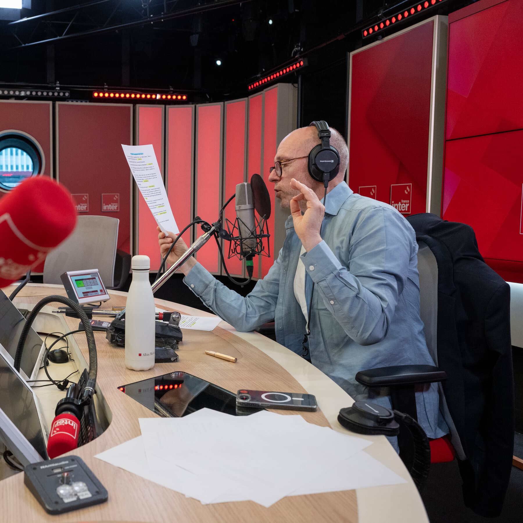 A satirical illustration in a moody, muted style. A classic French cafe scene with a single patron sitting at a round table. Instead of wine, they are staring at a vintage microphone on the table that is casting a long, dark shadow shaped like a World War II soldier or a general. The atmosphere is smoky and intellectual, with a sense of historical weight pressing down on the modern setting.