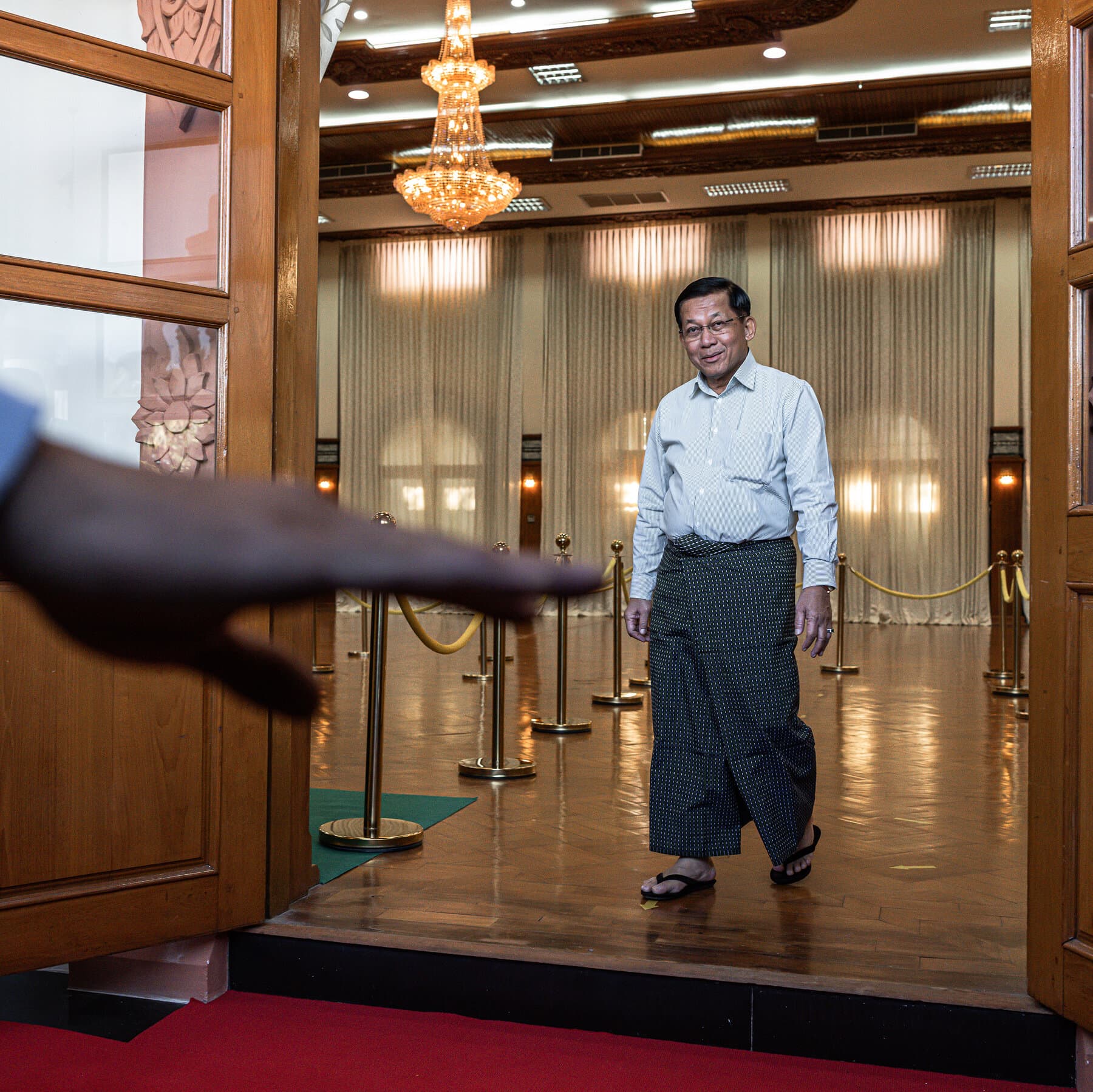 A gritty, editorial-style illustration of a stern military general in a blood-stained uniform, awkwardly trying to put on a shiny, oversized presidential sash while standing on a pile of gray rubble. The background is a dark, smoky sky. High contrast, cynical, and dark colors.
