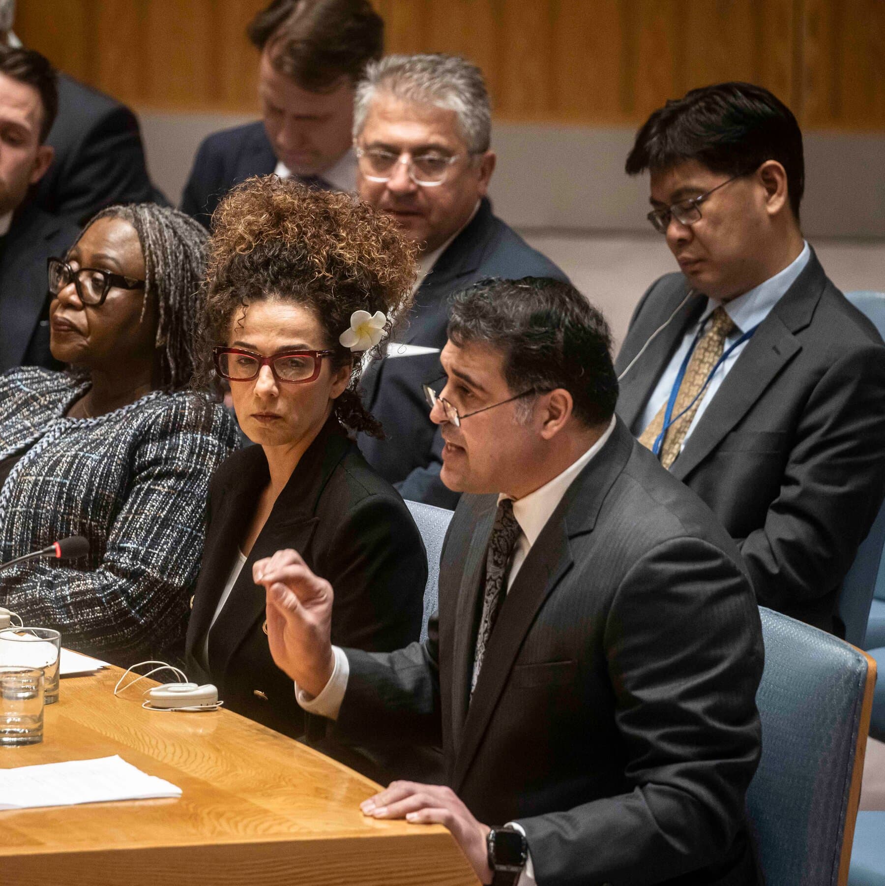 A photorealistic news image of a U.S. Ambassador at the UN Security Council pointing aggressively at a map of Iran while holding a large red button, while other diplomats look on with feigned concern.