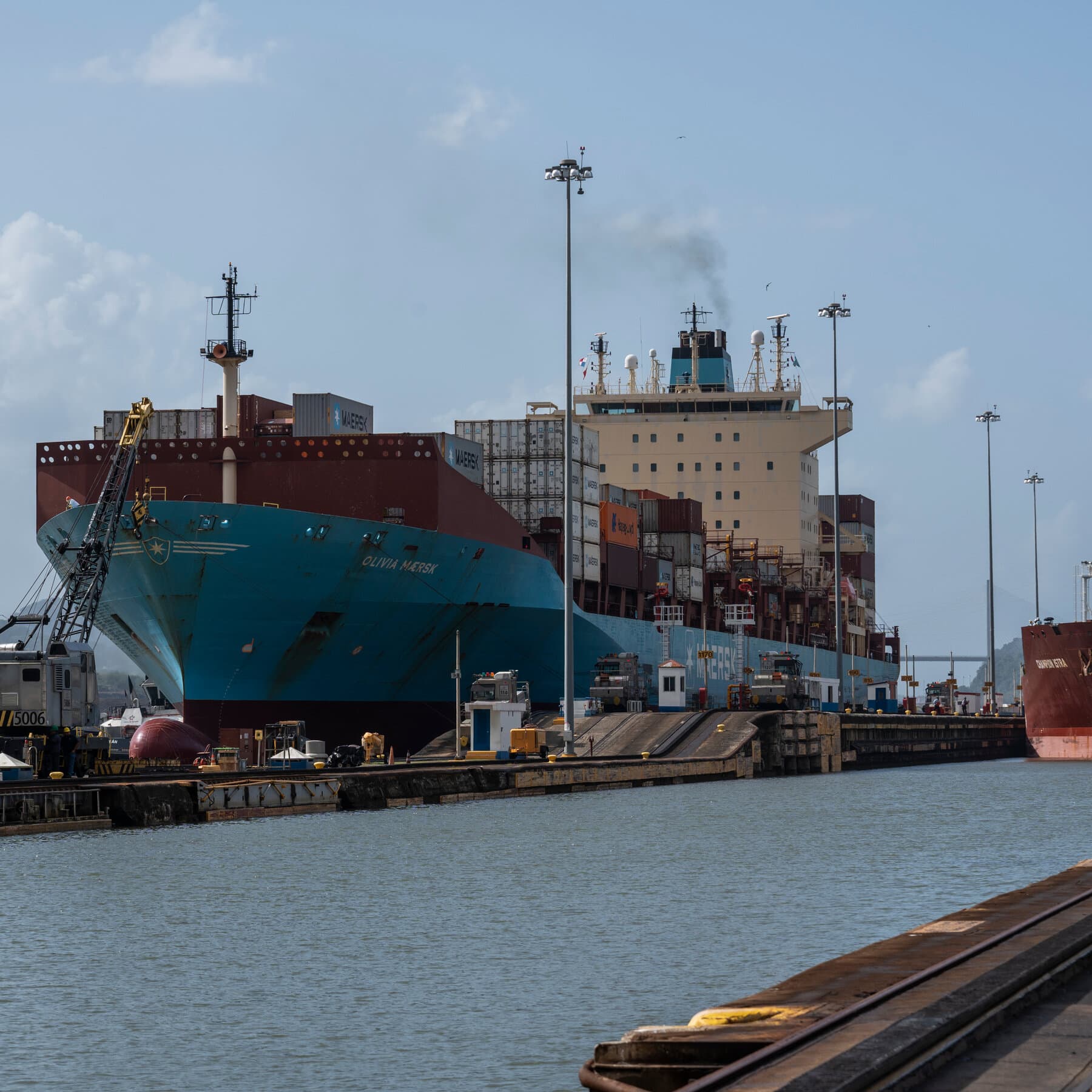 A moody, cynical editorial illustration showing a chessboard superimposed over a map of the Panama Canal. On one side, a large, shadowy hand places a US flag piece; on the other, a hand removes a piece shaped like a shipping container. The atmosphere is tense, misty, and colored in dull blues and greys, representing the cold reality of geopolitical power struggles.