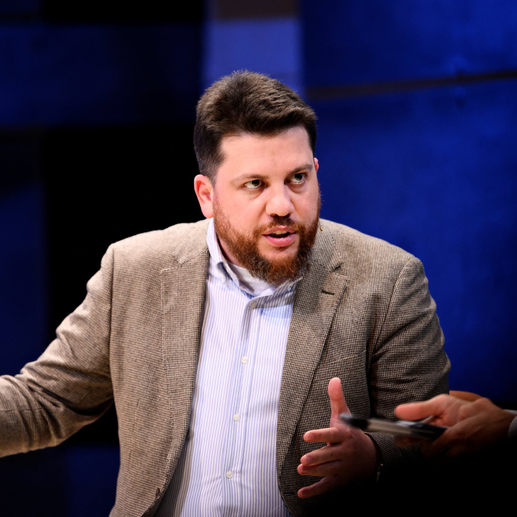 A sad-looking man with messy hair and round glasses sitting in a trendy European coffee shop wearing a shirt that says 'One Of The Good Ones' in Cyrillic. He is holding a lukewarm latte and looking confused while a group of people at the next table glare at him with intense hostility. Photorealistic style, news editorial photography.