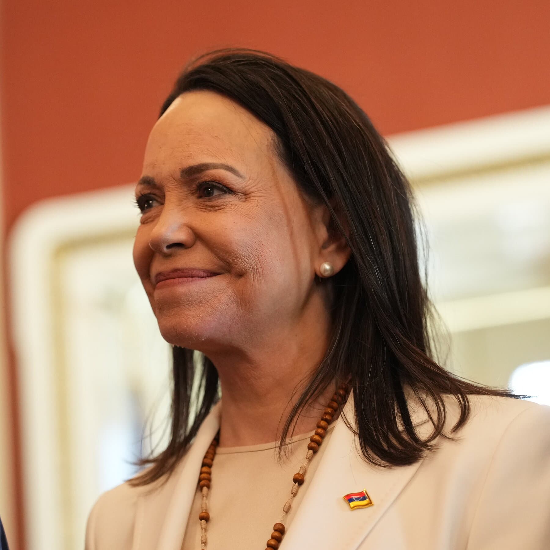 A photorealistic satirical image of Donald Trump sitting at a gilded table in Mar-a-Lago, wearing a tuxedo and squinting through a jeweler's loupe at a Nobel Peace Prize medal. He is holding a screwdriver in one hand, attempting to pry something off the medal. In the blurry background, a woman looking like María Corina Machado stands awkwardly by a velvet rope, holding an empty velvet box.