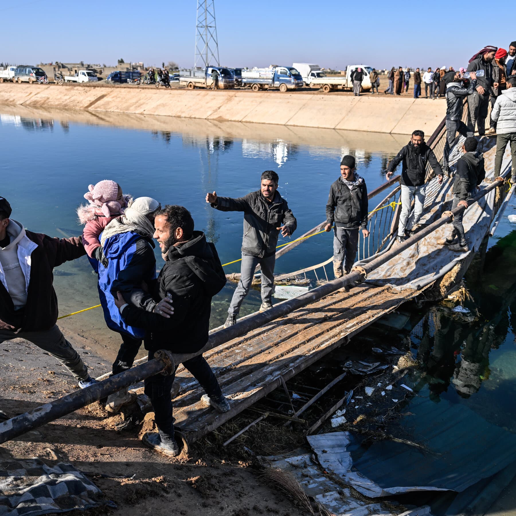 A satirical news photo of Syrian President Ahmed al-Sharaa standing at a podium giving a warm, welcoming thumbs-up gesture. Behind him, out of focus, a tank is actively blasting a hole through a brick wall. The caption style is high-contrast, realistic photojournalism.