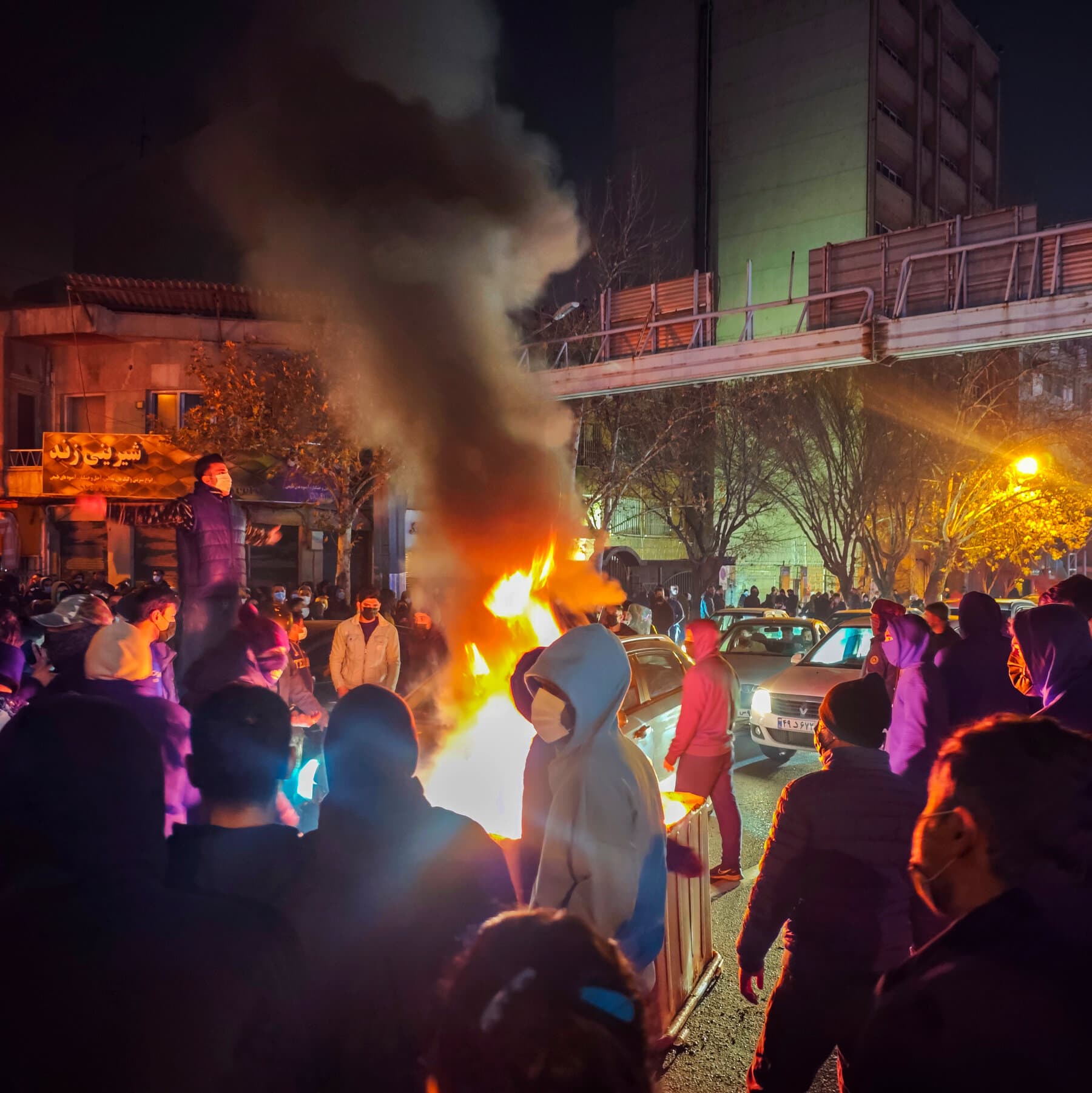 A serious-looking political analyst in a suit and glasses sitting in a cable news studio, pointing authoritatively at a map of Iran while a breaking news chyron reads 'IS THIS THE TIPPING POINT? (AGAIN)'.