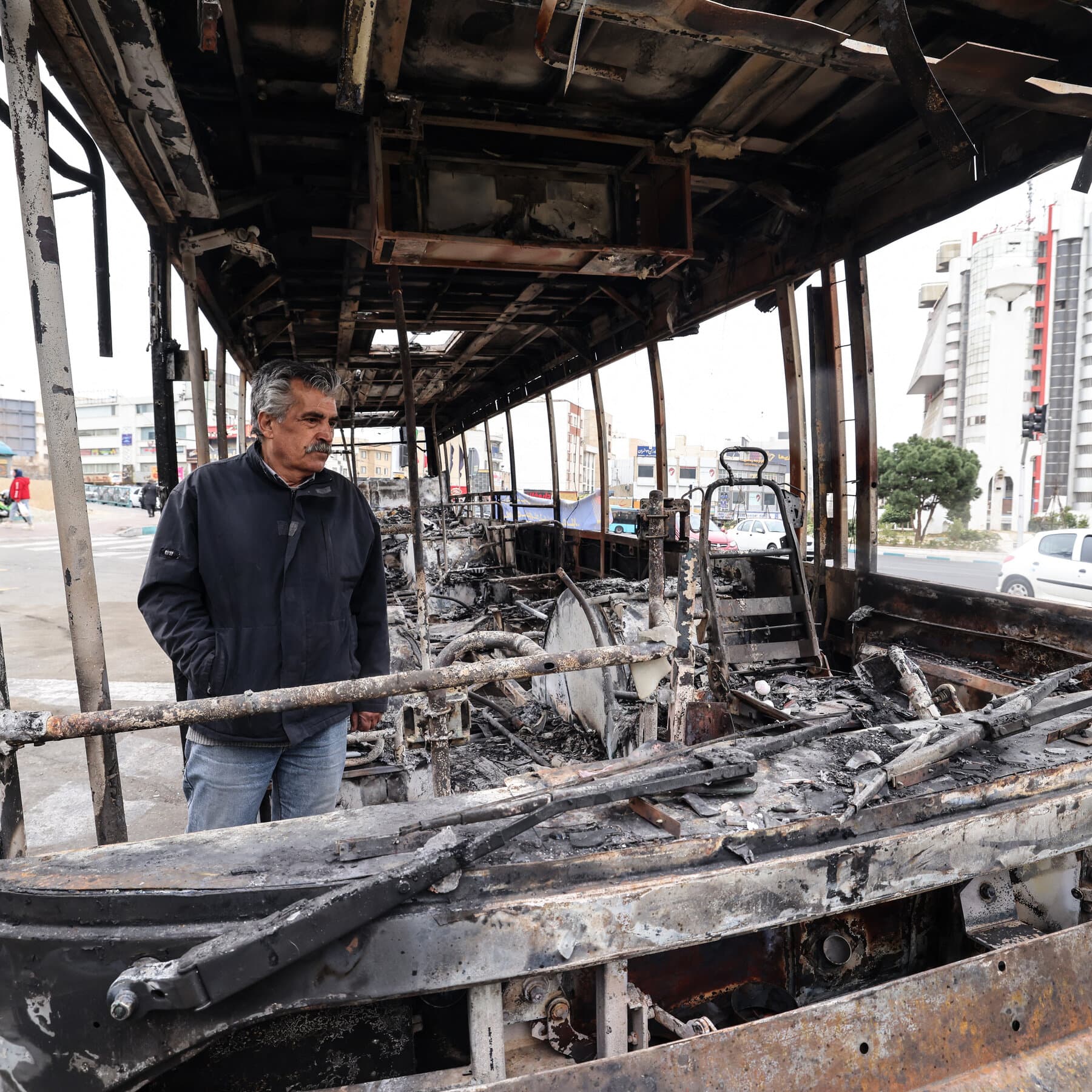 A satirical news photo featuring a high-ranking Iranian General standing at a podium labeled 'Mission Accomplished,' smiling broadly and gesturing to a large chart behind him that shows a line graph labeled 'Public Dissent' plummeting to zero, while outside the window behind him, a tank rolls down a completely empty, smoke-filled street.