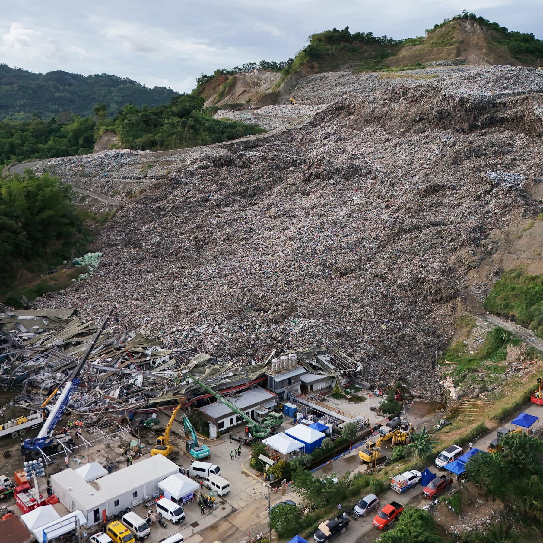 A satirical news photo style image. A serious government official in a suit and yellow hard hat stands at a podium with microphones in the foreground. In the background is a massive, chaotic mountain of garbage. The official is holding up a blueprint that clearly shows a diagram of a pile of trash. The mood is somber but absurd.