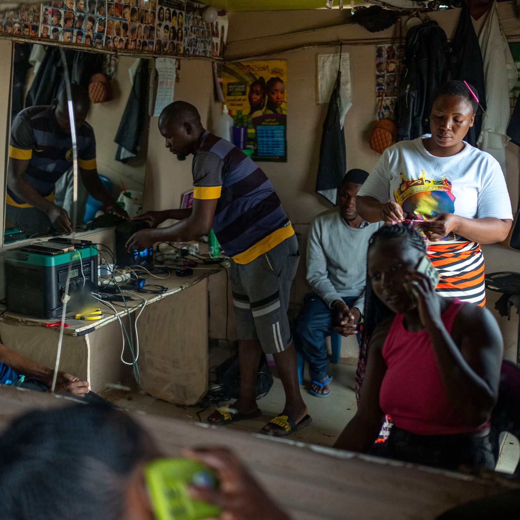 A photojournalistic image of a smiling man in a dark room proudly holding a glowing, bulky car battery like it is a newborn baby, while his family gathers around to warm their hands over the gentle hum of an LED indicator light.