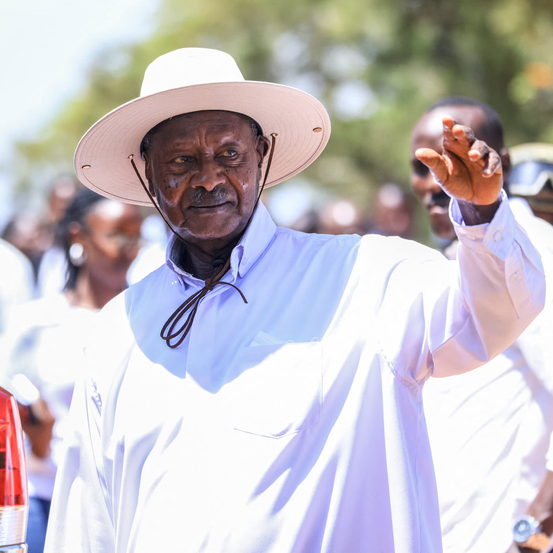 A satirical photo-realistic image of Ugandan President Yoweri Museveni wearing his signature wide-brimmed hat, looking feignedly surprised and holding a hand over his mouth while standing next to a ballot box that is overflowing with ballots clearly written in the same handwriting, confetti falling in a lavish palace room.