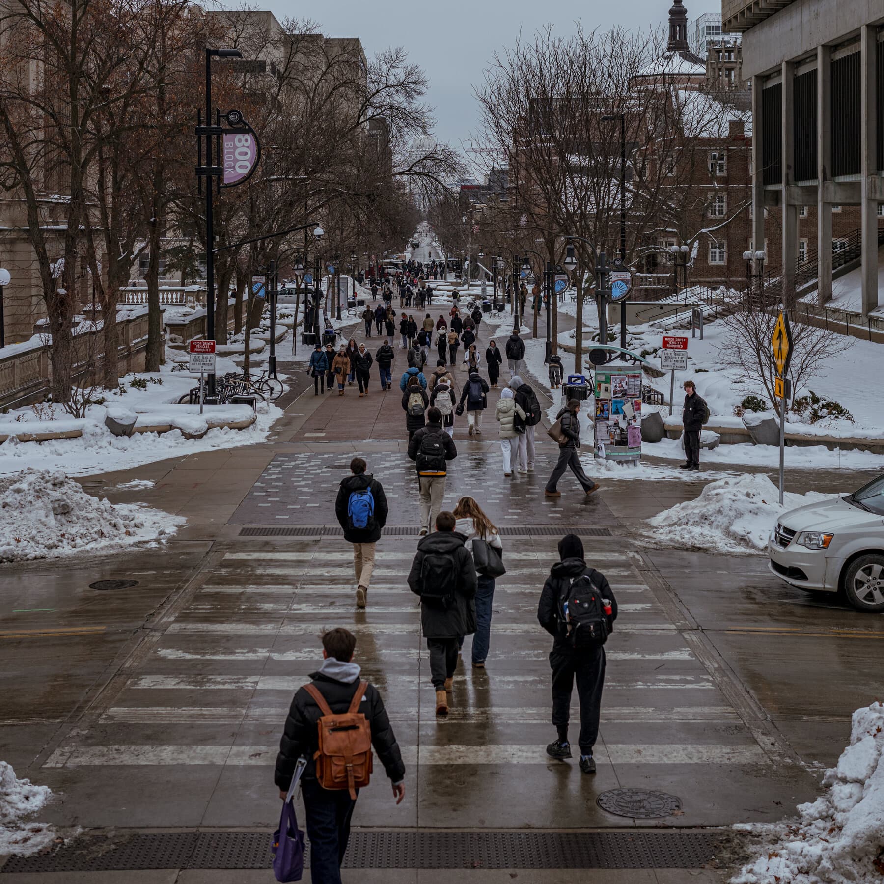 A desolate, brutalist university campus in winter, snow covering empty benches. In the foreground, a torn 'Welcome' banner flutters in the wind. The atmosphere is grey, cold, and abandoned, symbolizing economic and intellectual emptiness.