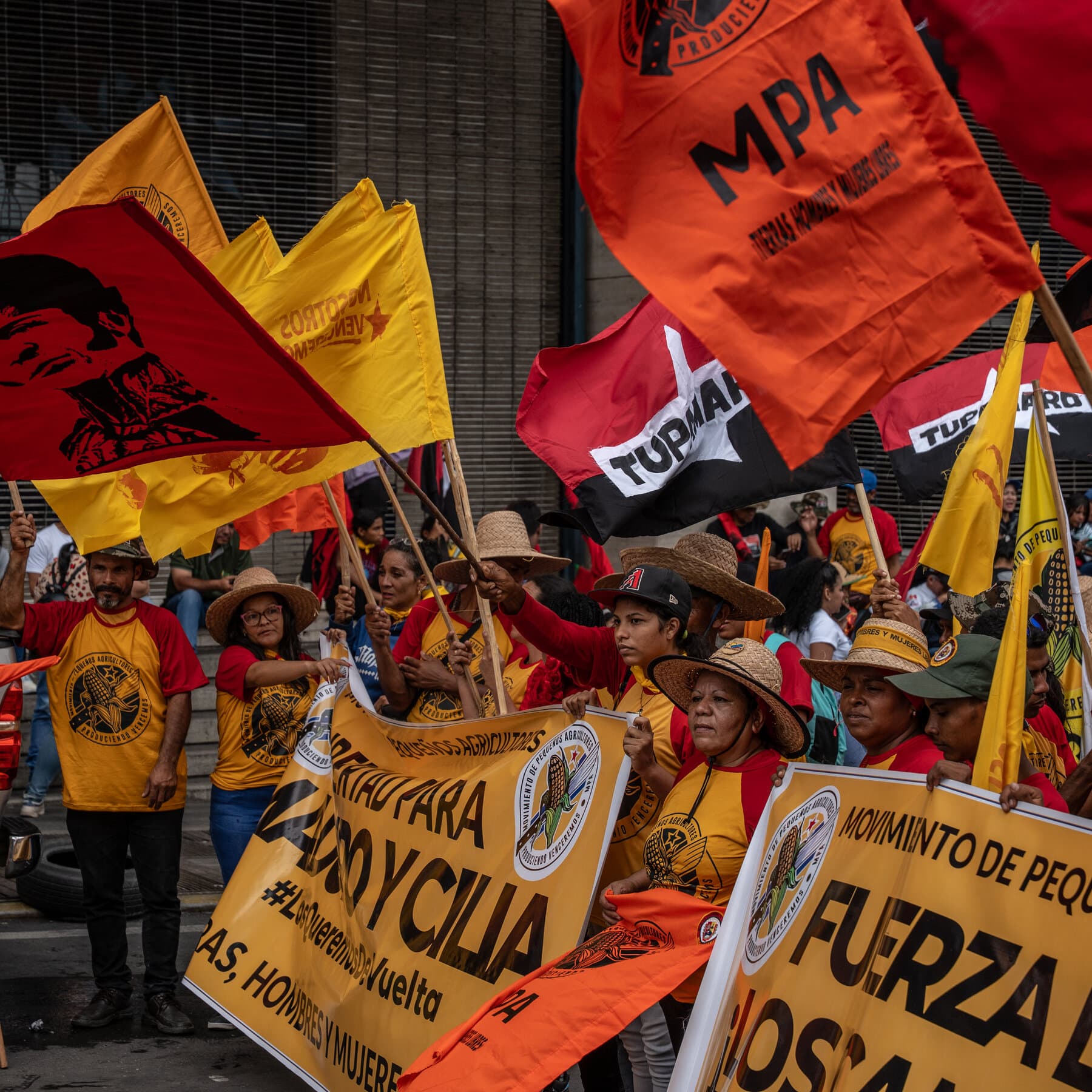 A cynical oil painting style illustration showing a tattered, faded red Venezuelan 'Bolivarian' flag being stitched together with a pristine US dollar bill. In the background, a silhouette of a leader in a red beret shakes hands with a faceless bureaucrat in a suit, both standing on a pile of empty oil barrels in a desolate landscape. The lighting is dim, dusty, and melancholic.