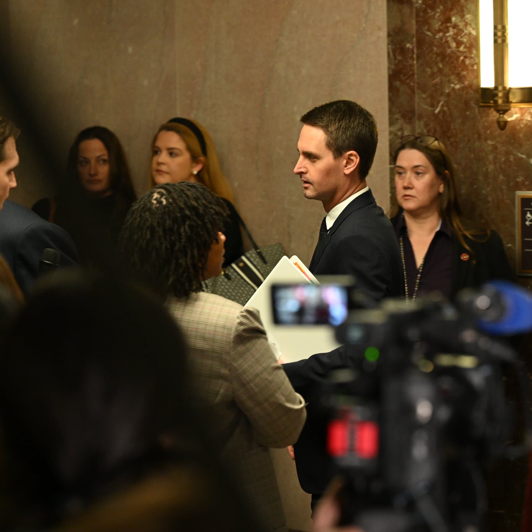 A satirical, dark-humored illustration of a yellow Snapchat ghost wearing a business suit, holding a massive briefcase overflowing with cash, standing atop a pile of discarded, glowing smartphones in a courtroom. The style is acid-etched political cartoonery, high contrast, with a cynical and grim atmosphere.