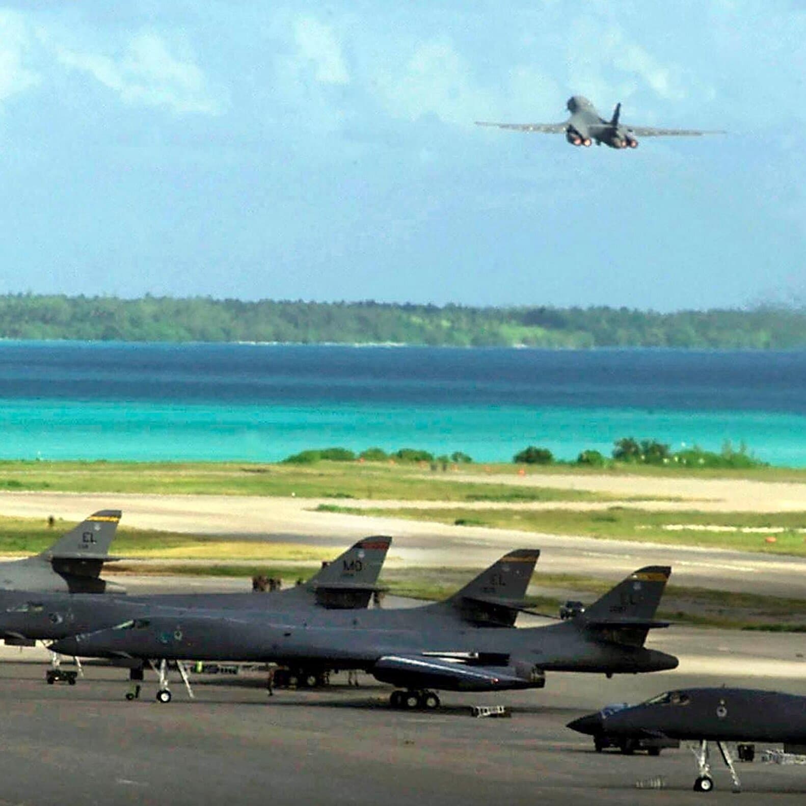 A satirical news photo showing a British diplomat handing a Mauritian official a single grain of sand while standing in front of a massive, barbed-wire fence guarding a stealth bomber on a tropical beach.