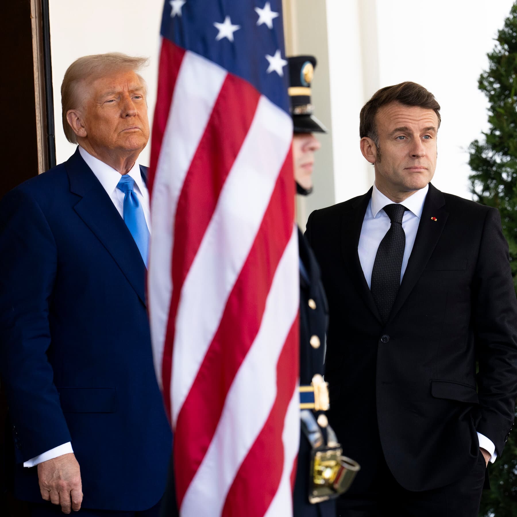 A photorealistic news style image of French President Emmanuel Macron sitting awkwardly on Donald Trump's lap in the Oval Office. Macron looks up with wide, pleading puppy-dog eyes while Trump looks absentmindedly at a TV screen, resting one hand heavily on Macron's head like he is a golden retriever. High contrast, professional lighting.